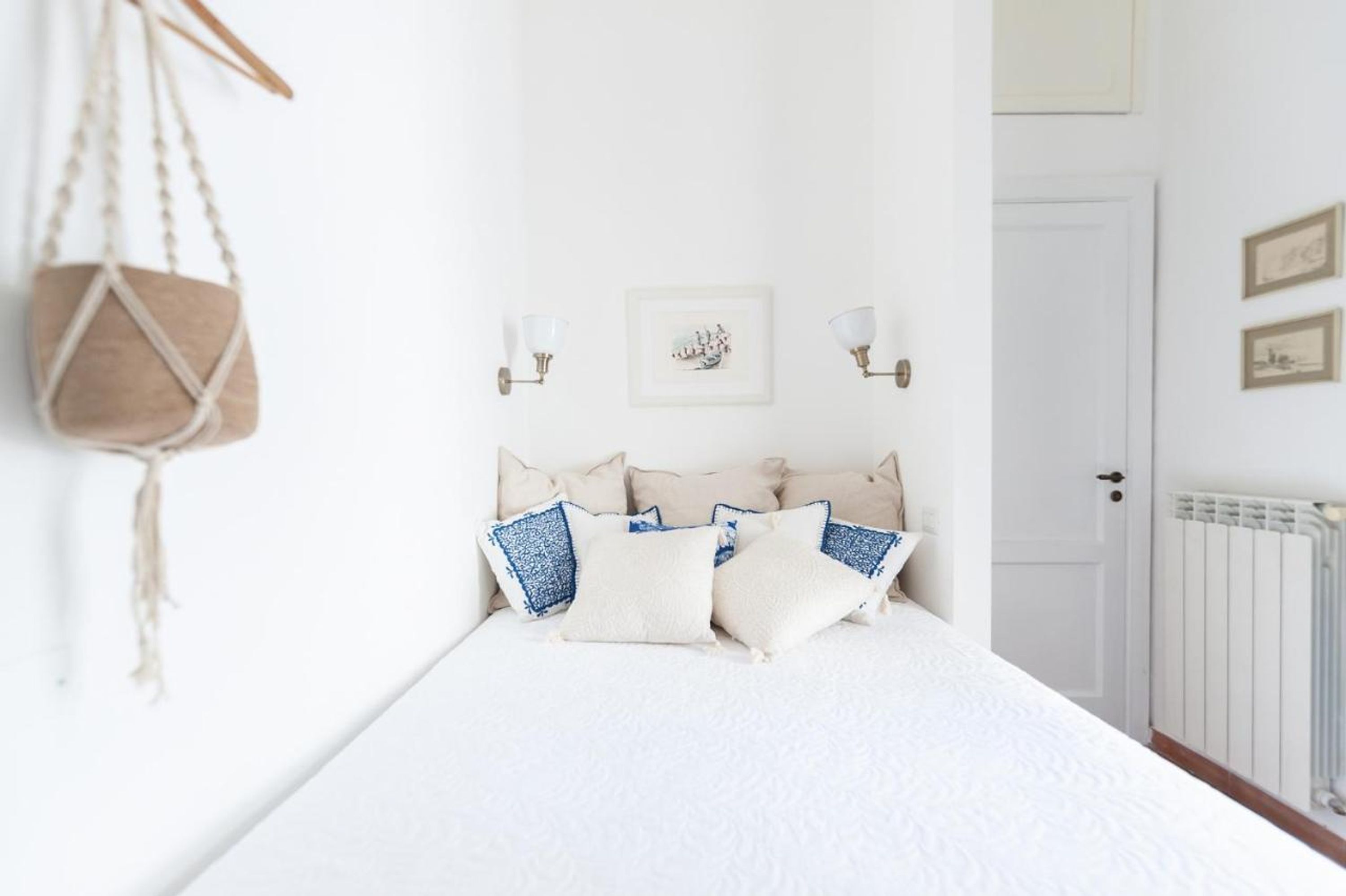 A neatly made bed with a white duvet and several pillows, a wall-mounted light fixture, and a framed picture on the wall.