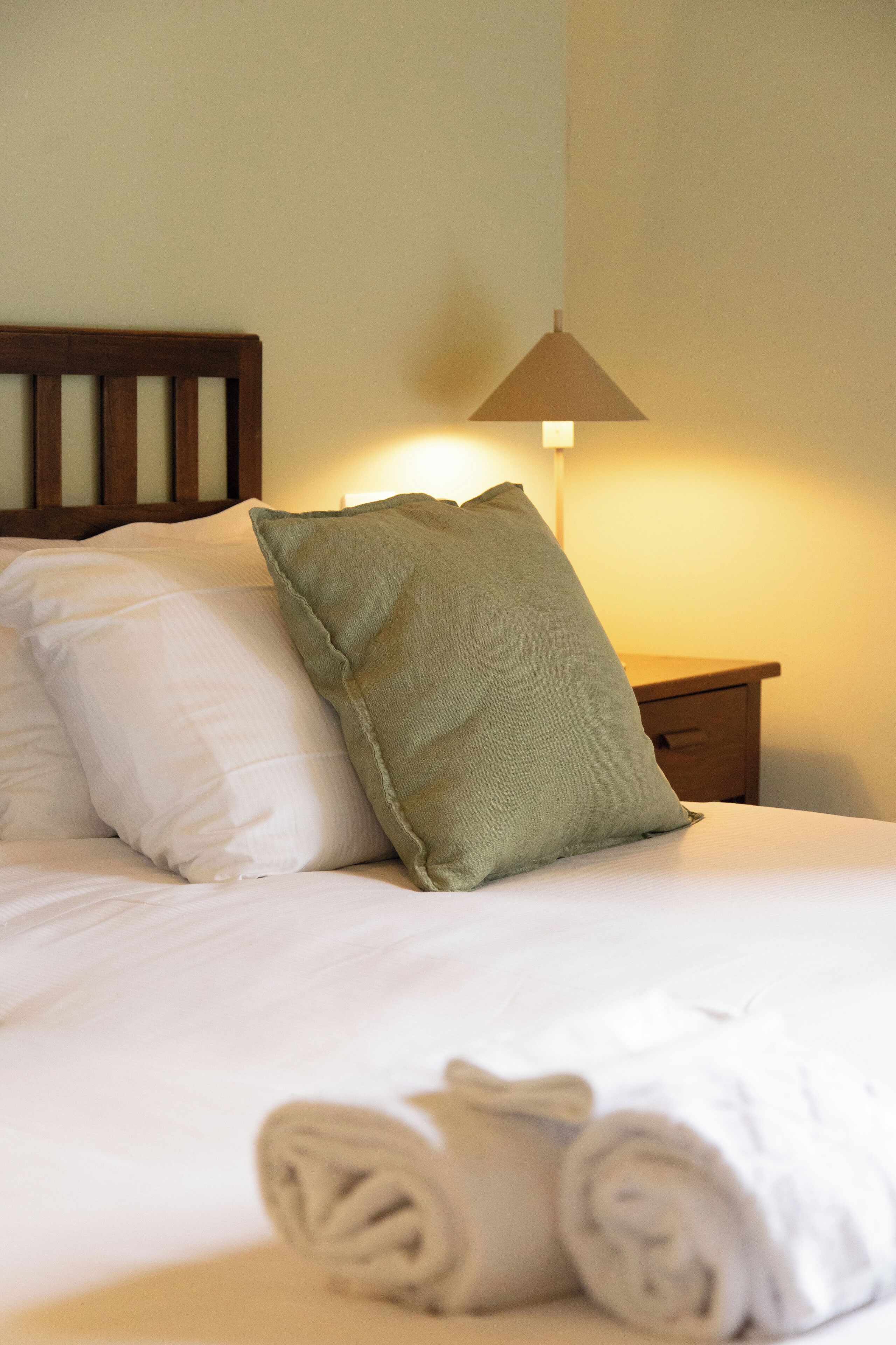 Uma cama bem arrumada com roupa de cama branca e um travesseiro verde, uma cabeceira de madeira e uma mesinha de cabeceira com uma luminária.
