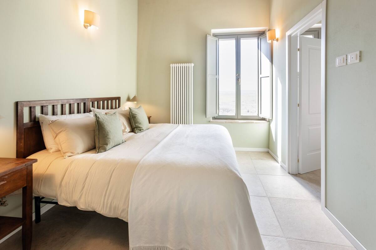 Quarto com cama, mesa de cabeceira, abajur, radiador, janela e porta.