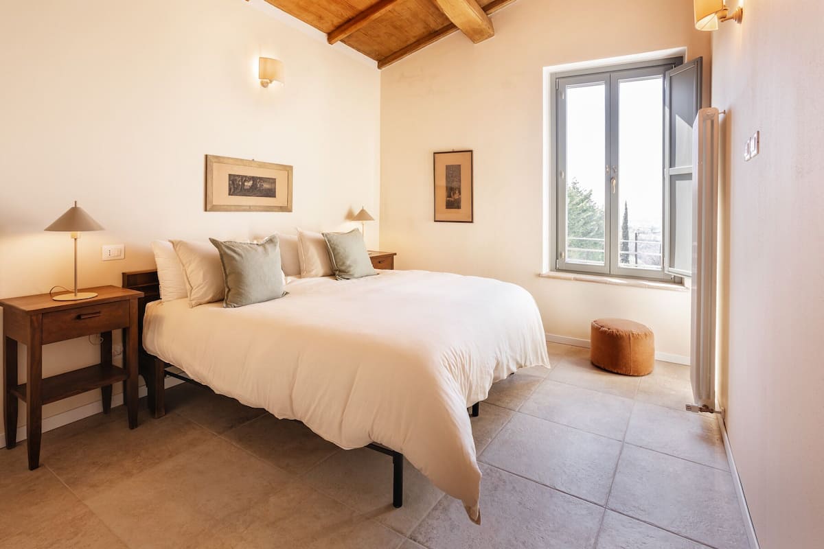 Quarto com uma cama grande, duas mesas de cabeceira, uma janela com vista e duas luminárias de parede.