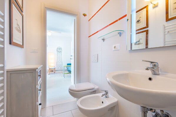 Une salle de bain moderne avec un grand lavabo, un bidet et des toilettes. Les murs sont ornés de tableaux encadrés et une étagère en verre y est fixée.