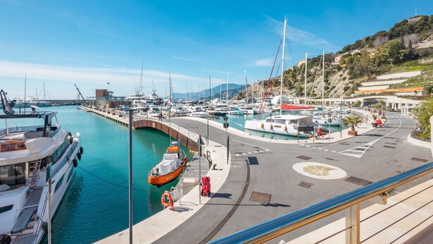 Un port de plaisance où les bateaux sont amarrés le long d’une promenade en bord de mer aux courbes élégantes, avec un sentier piétonnier et une vue sur des collines en arrière-plan.