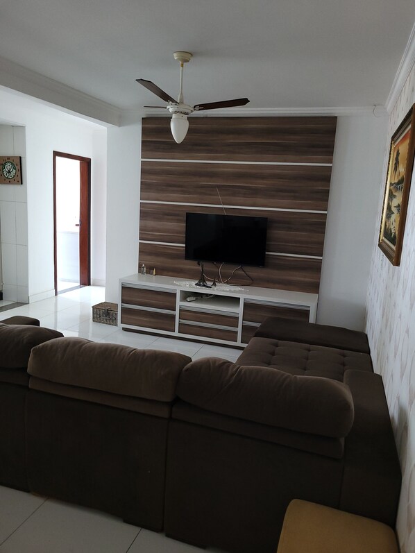 Una sala moderna con un amplio sofá modular, un televisor de pantalla plana montado en una pared de paneles de madera, un ventilador de techo y una puerta que da a otra habitación.