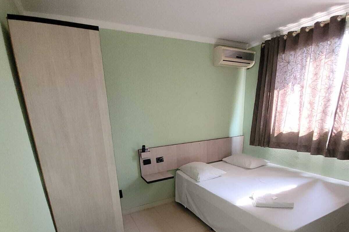 Habitación individual con una cama, un armario de madera, una mesita de noche pequeña y una ventana con cortinas.