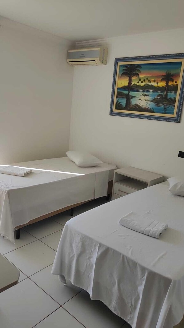 A hotel room with two single beds, a nightstand, a wall-mounted air conditioner, and a tropical landscape painting. - Hotel Globo de Cianorte (Cianorte)