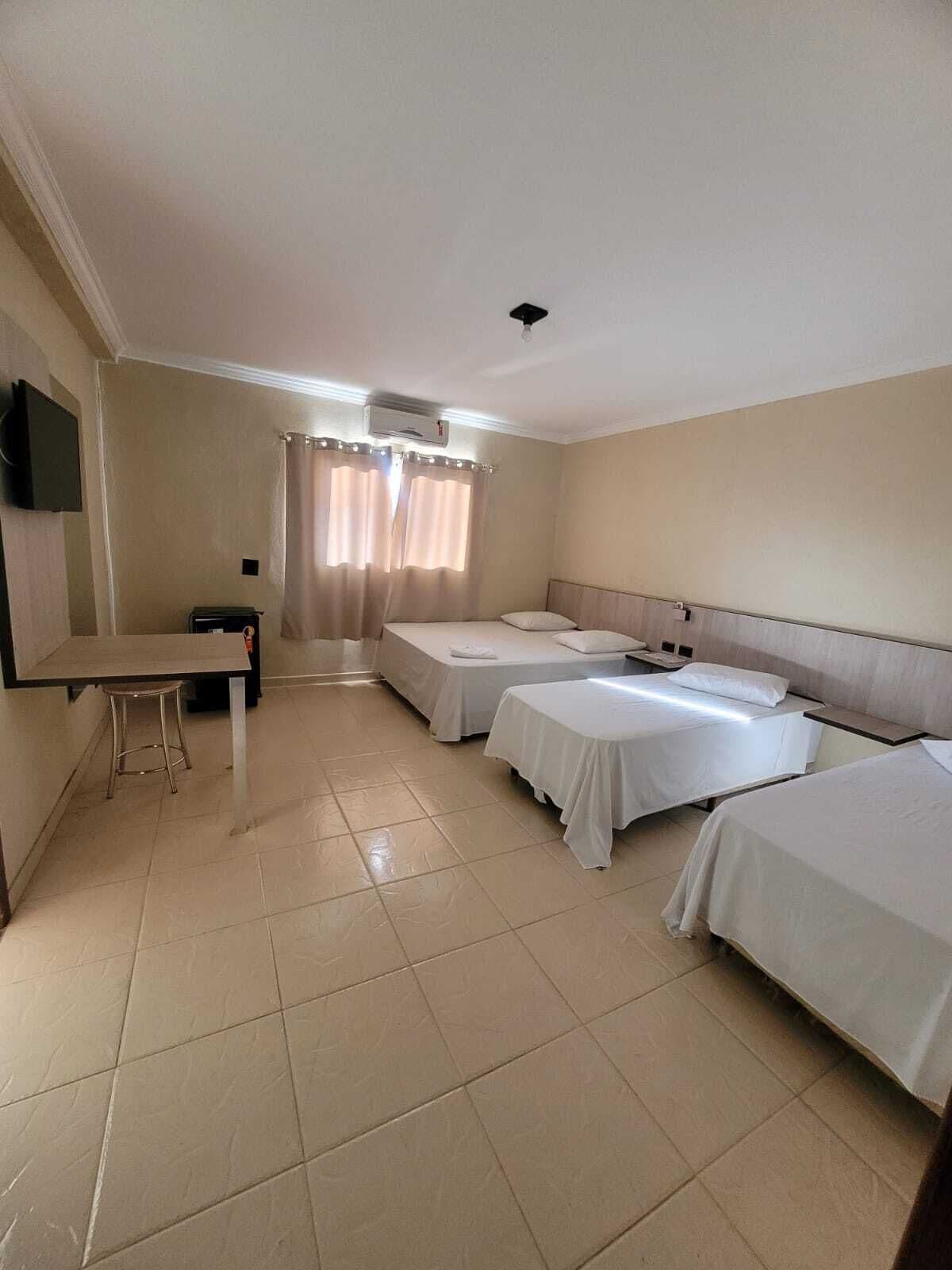 A hotel room with two single beds, a small desk with a chair, a TV mounted on the wall, and a window with curtains.