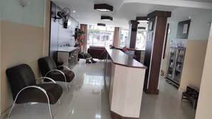 Zona de recepción de hotel con mostrador, varias sillas, un ventilador y un refrigerador.