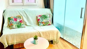 Une pièce accueillante avec un canapé, deux coussins, une petite table ornée d’une plante et une porte-fenêtre coulissante donnant sur un espace extérieur.