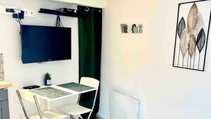 Une petite chambre moderne avec un téléviseur à écran plat fixé au mur, une table pliante avec deux chaises, une petite plante en pot et une œuvre d’art encadrée accrochée au mur.