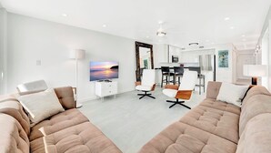 Ett modernt vardagsrum med en stor platt-TV, två soffor, en matplats med stolar och ett kök i bakgrunden.