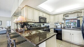 A spacious kitchen with white cabinetry, a granite countertop, and a central island with a lamp. - Indies West at The Moorings (Naples)