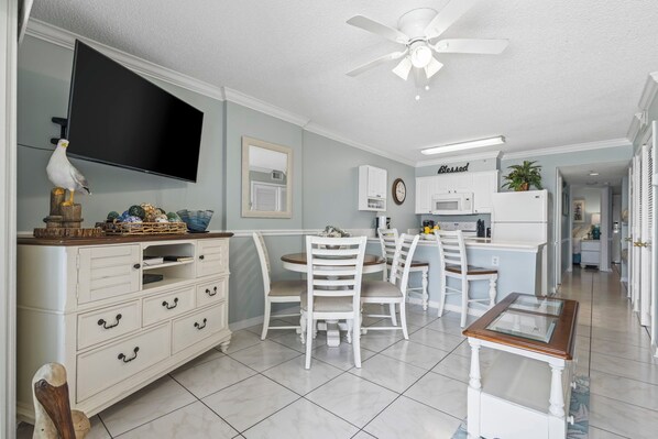 A kitchen with white cabinetry, a dining table with chairs, a ceiling fan, and a television mounted on the wall.