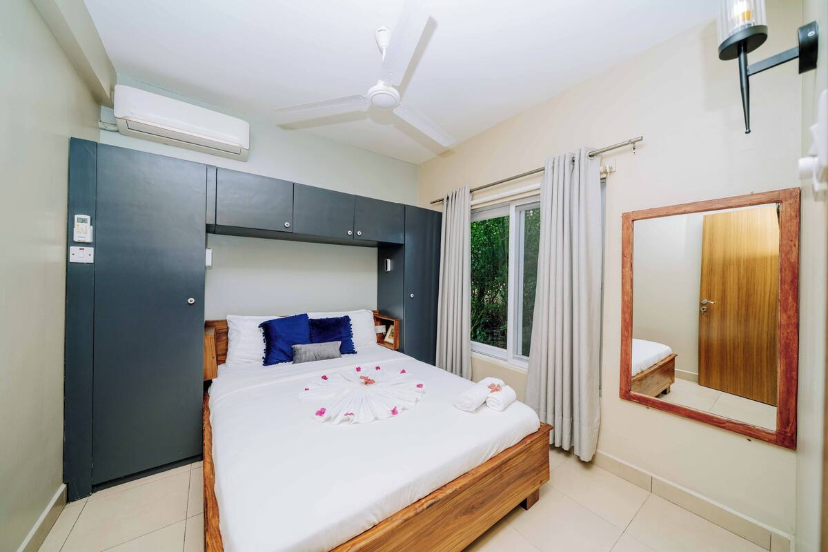 Une chambre à coucher comprenant un lit, un ventilateur de plafond, un climatiseur, une fenêtre avec des rideaux, une armoire en bois et un miroir.