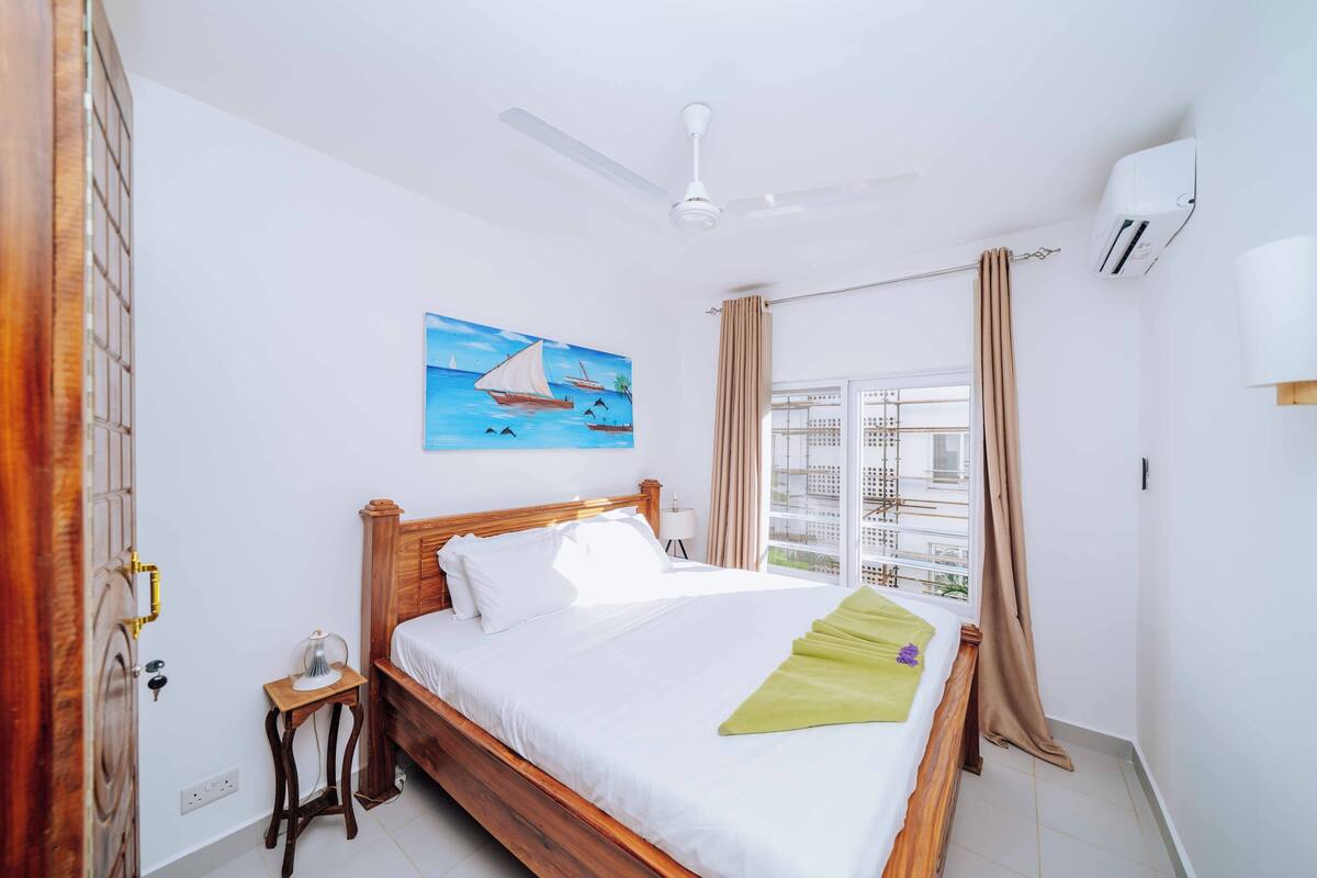 Une chambre à coucher avec un lit en bois, un ventilateur de plafond, une fenêtre avec des rideaux et un tableau accroché au mur.
