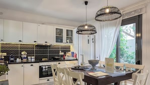 A modern kitchen with white cabinets, a black countertop, and a dining area with white chairs. - Alisahni Villa (Daratsos)