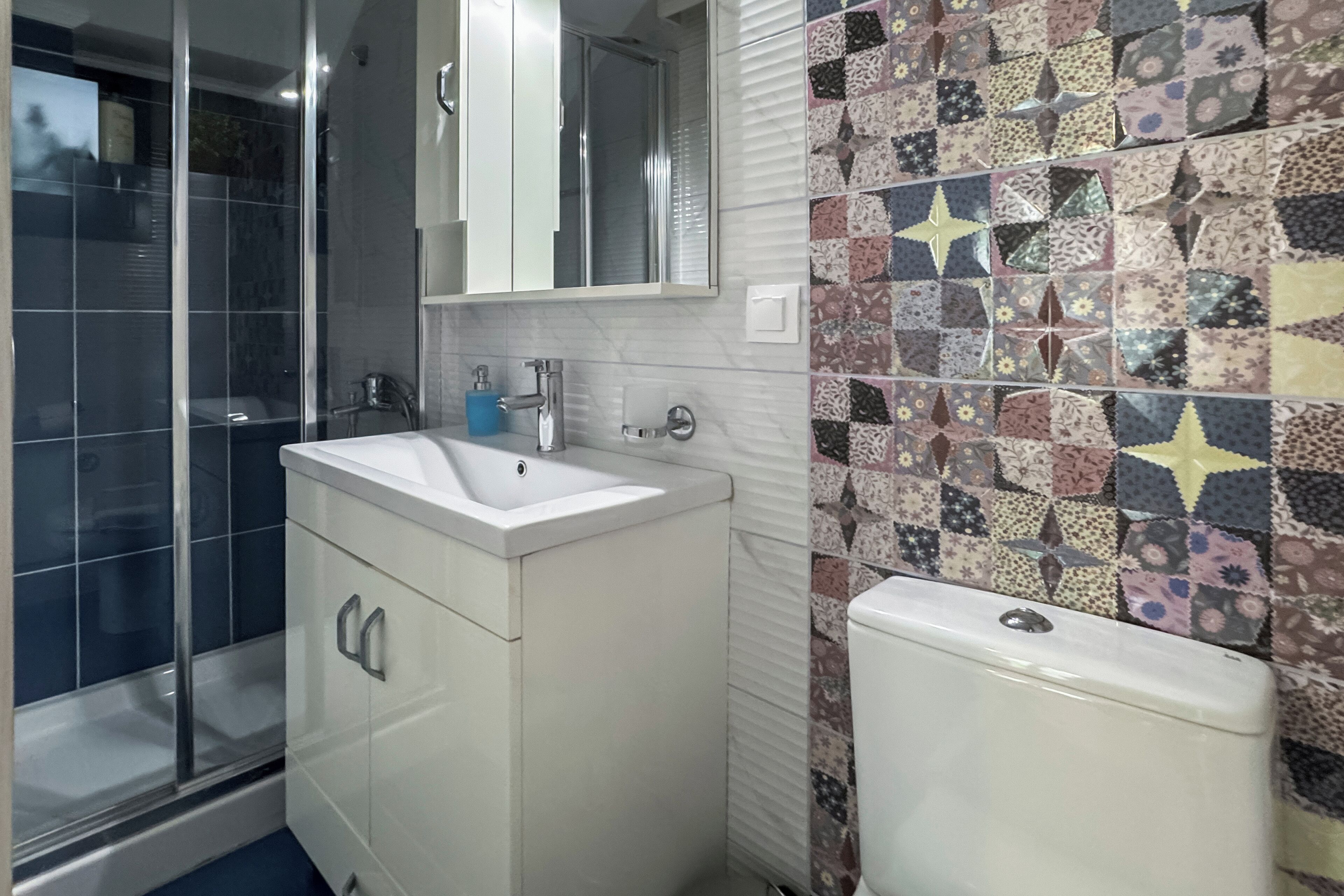 Ein Badezimmer mit weißem Waschbecken, einem Spiegel darüber, einer Duschkabine und einer Toilette. Die Wände sind mit verschiedenen Mustern und Farben gefliest.