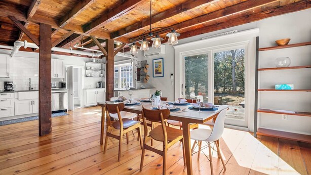 A dining area with a wooden table and chairs, a kitchen in the background, and a view of the outdoors through large windows. - Steps to Dining, Less than 1 mile to Bayside Beaches! (Dennis)