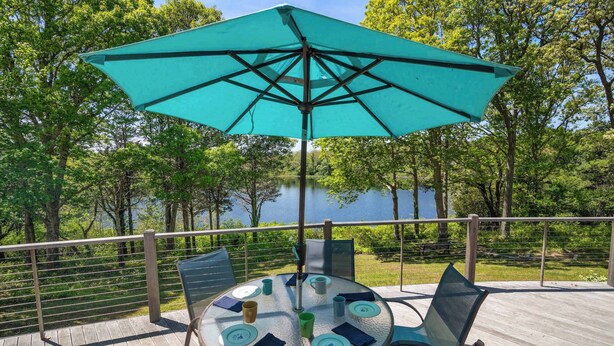 Een ronde tafel met stoelen en een turquoise parasol, met uitzicht op een meer met bomen op de achtergrond.