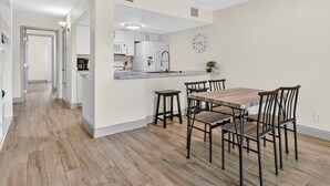 Una cocina moderna con electrodomésticos blancos, un comedor con mesa de madera y sillas metálicas, y un pasillo que conduce a otra habitación.