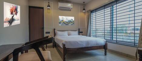 A hotel room with a bed, a desk, a chair, a ceiling fan, and a window with blinds.