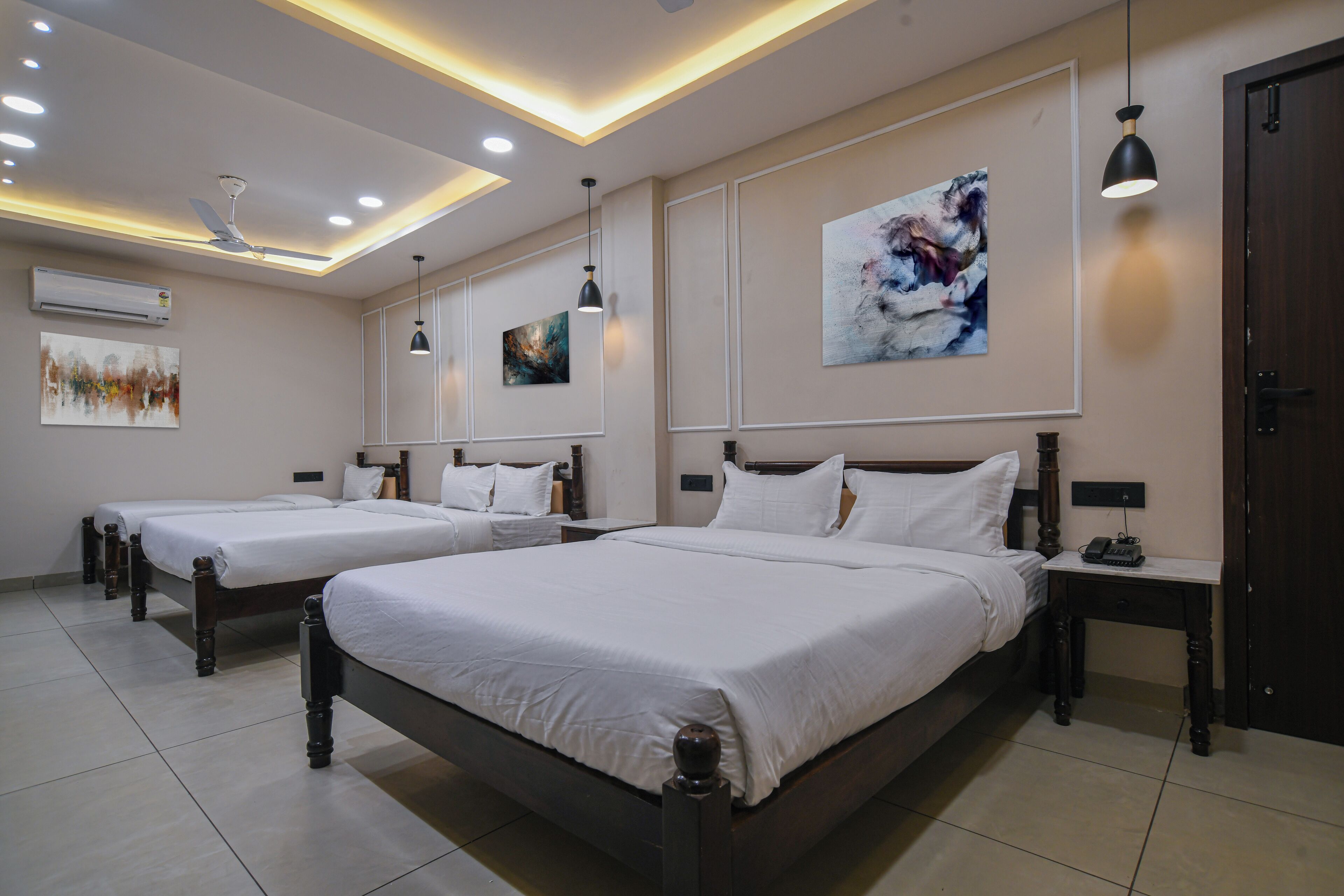 A hotel room with three beds, a ceiling fan, recessed lighting, artwork, and a bedside table with a lamp.
