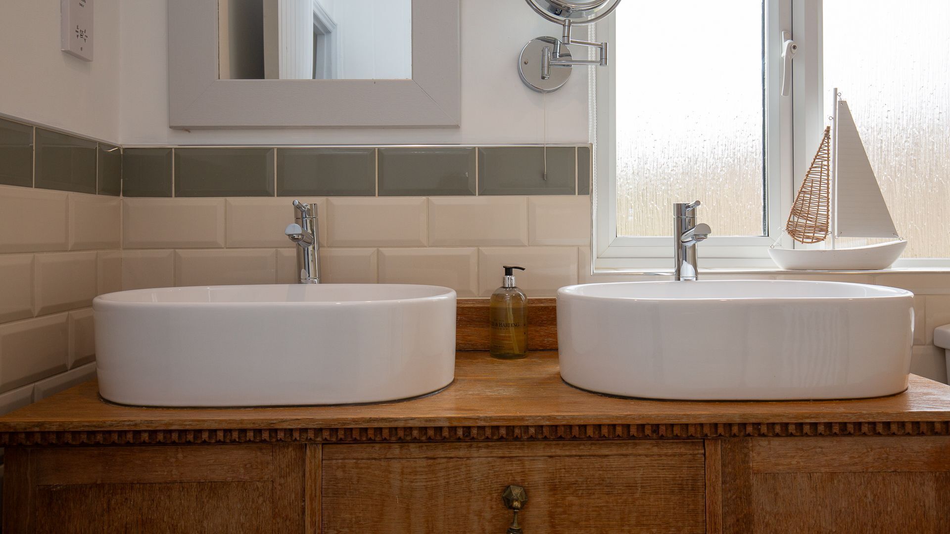 A bathroom with two white sinks, a wooden vanity, a mirror, and a window with a sailboat decoration.
