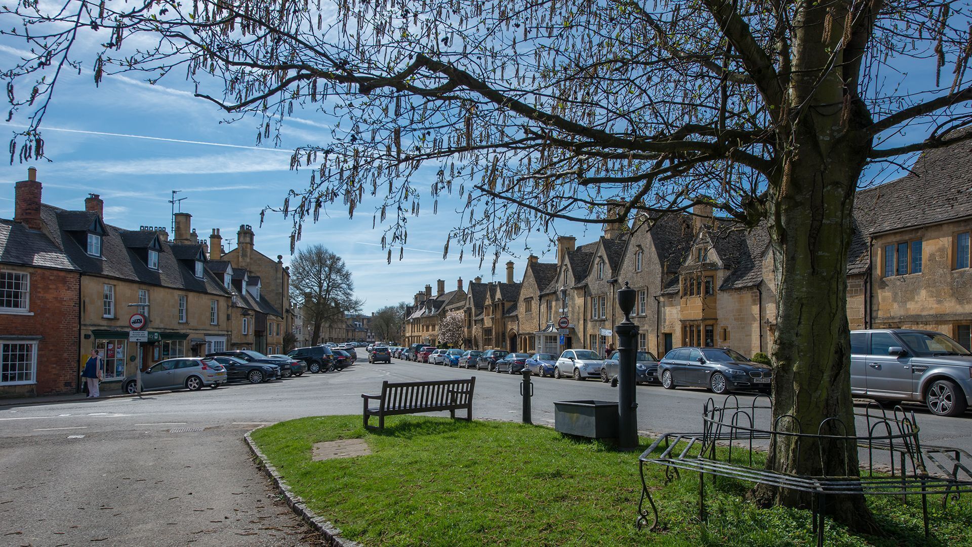 A street lined with traditional stone buildings, parked cars, and a bench in a grassy area.
