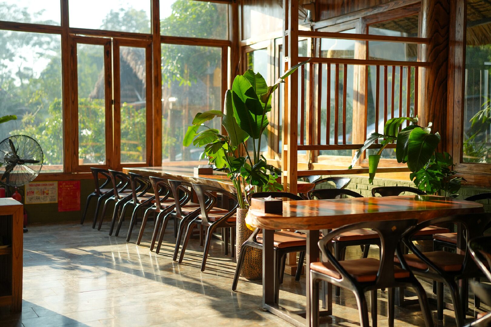 A spacious indoor area with large windows, wooden furniture, and potted plants.