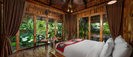 A large bed with a red and green bedspread, wooden flooring, and large windows with curtains.