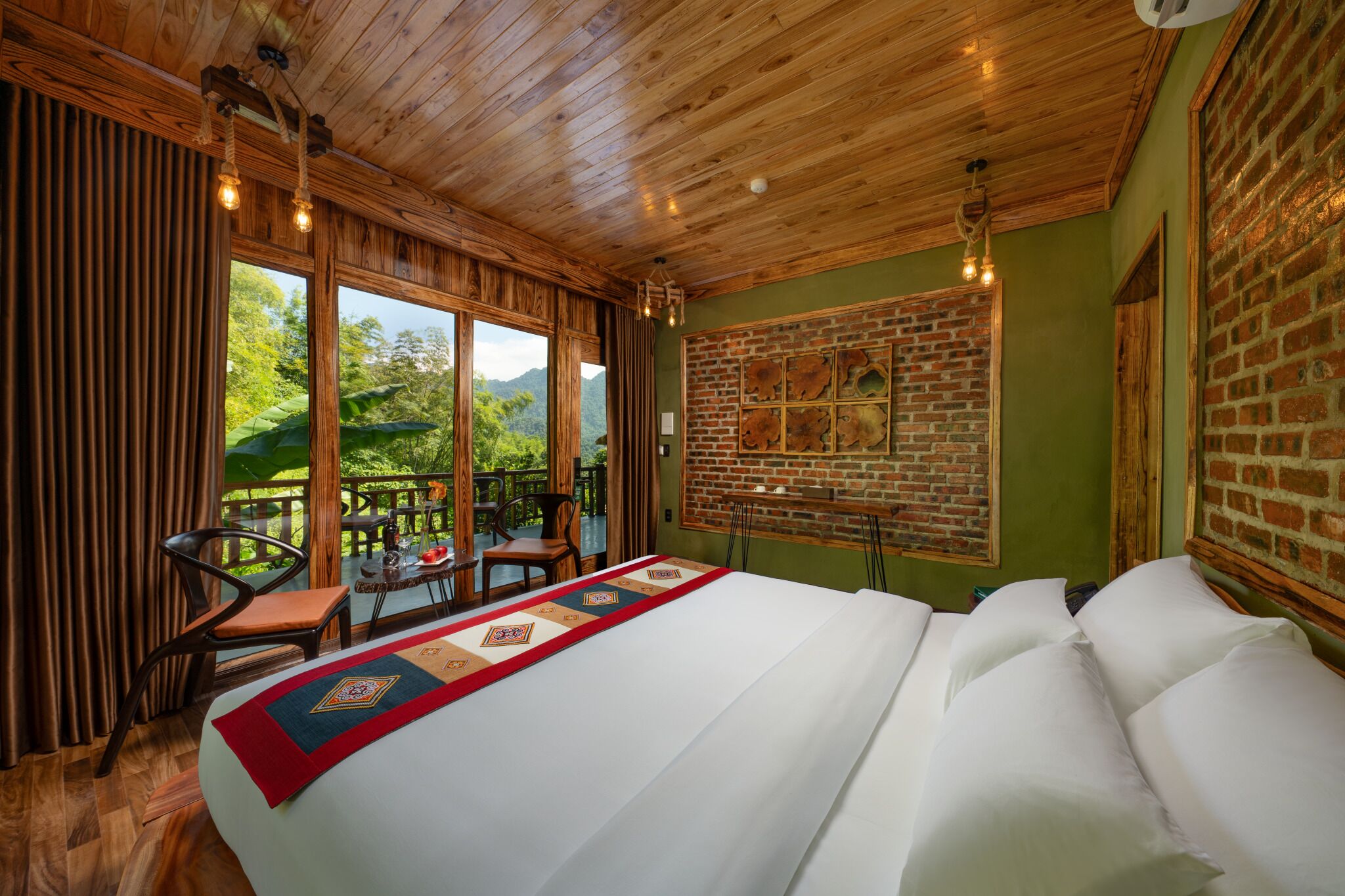 A hotel room with a large bed, wooden ceiling, a brick wall, and a view of greenery.