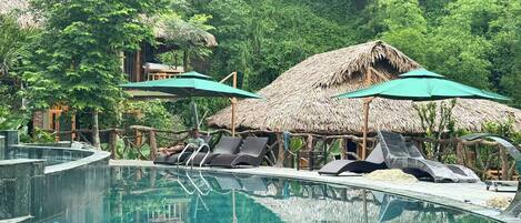 Zona de piscina con sillones de descanso y sombrillas, rodeada de vegetación exuberante y estructuras con techo de paja.