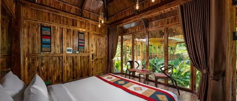 A wooden room with a bed, a table with chairs, and a view of greenery outside.
