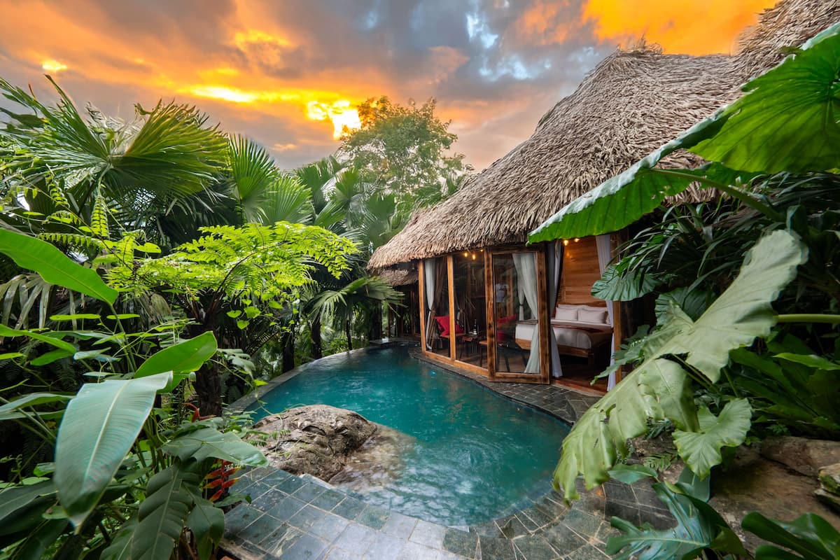 Zona junto a la piscina con una estructura de techo de paja, rodeada de exuberante vegetación tropical.