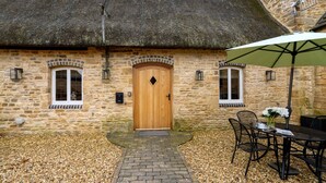 Une petite maison en pierre avec une porte en bois, deux fenêtres avec volets, un toit de chaume et un chemin de gravier menant à une table et des chaises, le tout abrité par un parasol.