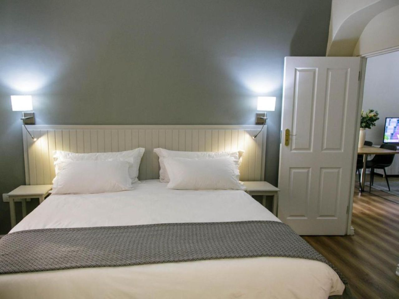 A bed with white linens, two bedside tables, a desk with a computer, and a door.