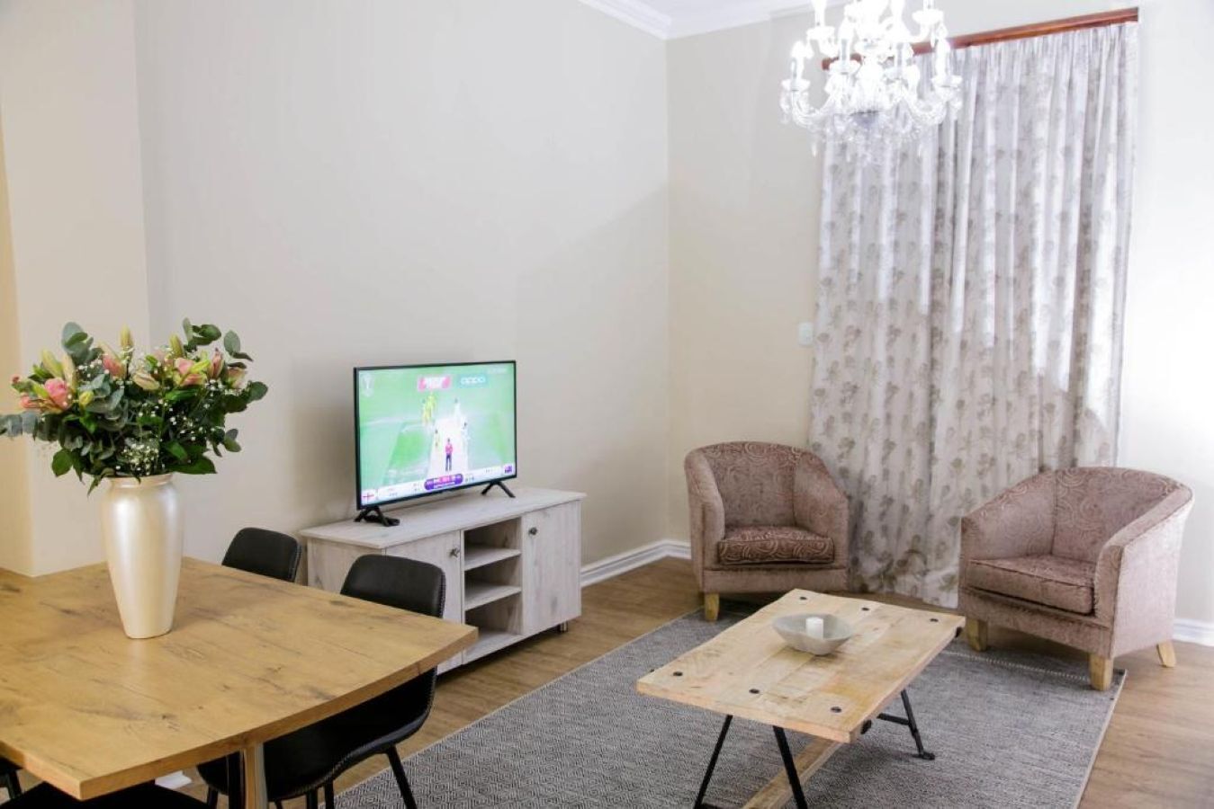 A living room with a TV, two armchairs, a wooden coffee table, a vase with flowers, and a chandelier.
