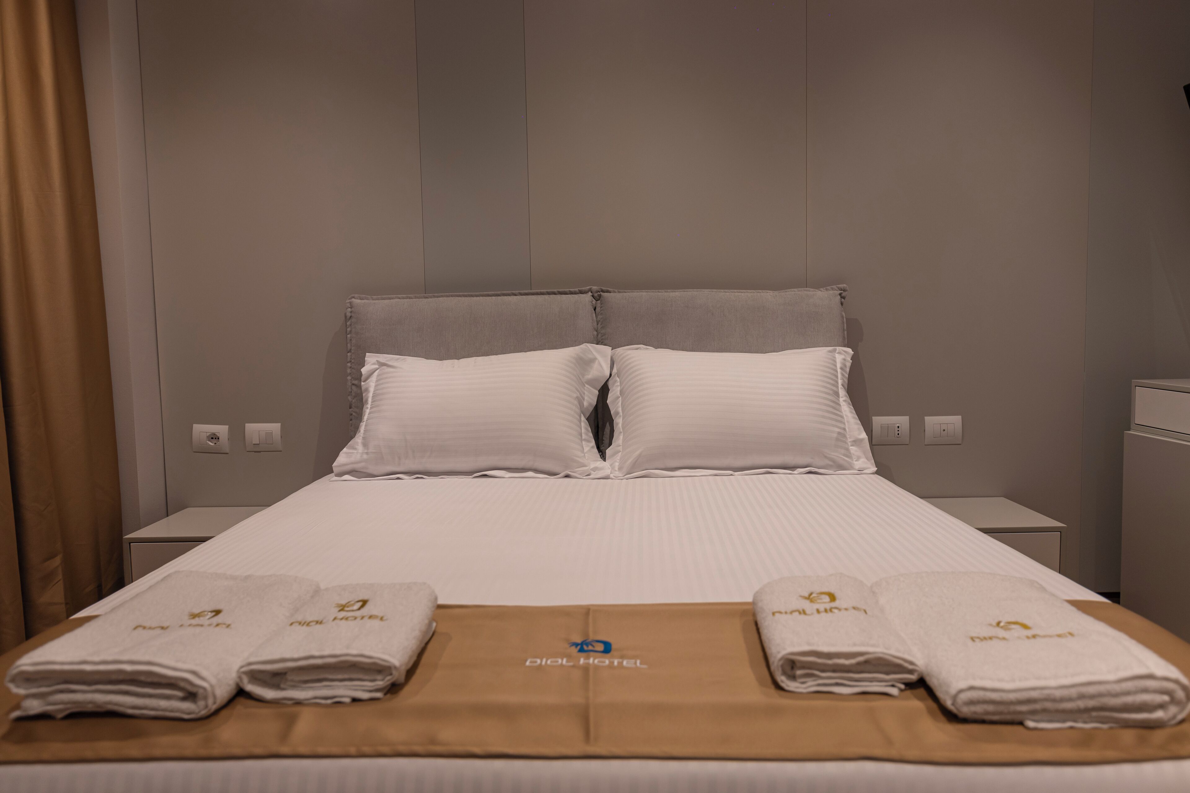 Habitación de hotel con una cama, dos almohadas, dos mesitas de noche y dos toallas con el logotipo del hotel.
