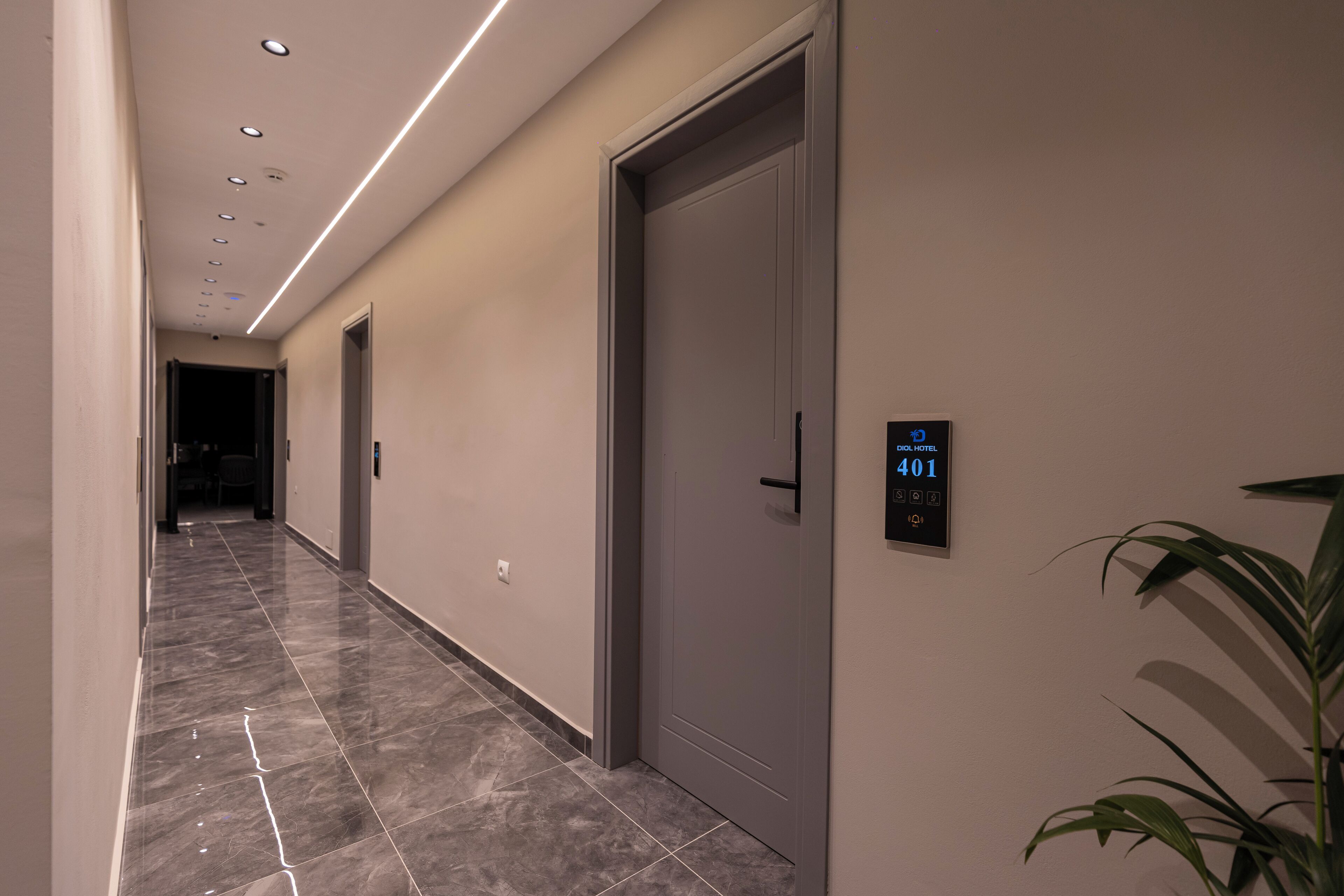 Un couloir d’hôtel moderne avec un sol en marbre, une porte dotée d’un écran numérique et une plante en pot.