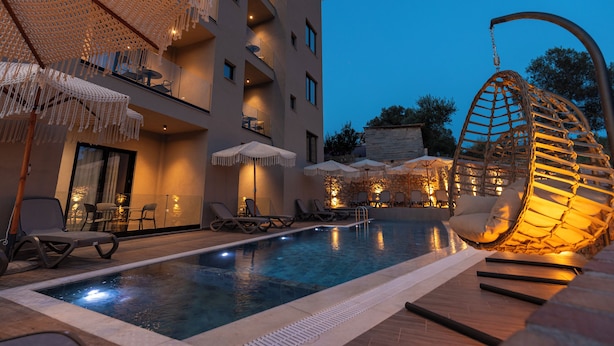 Un espace piscine avec des chaises longues et des parasols, un fauteuil suspendu et un bâtiment doté de balcons.