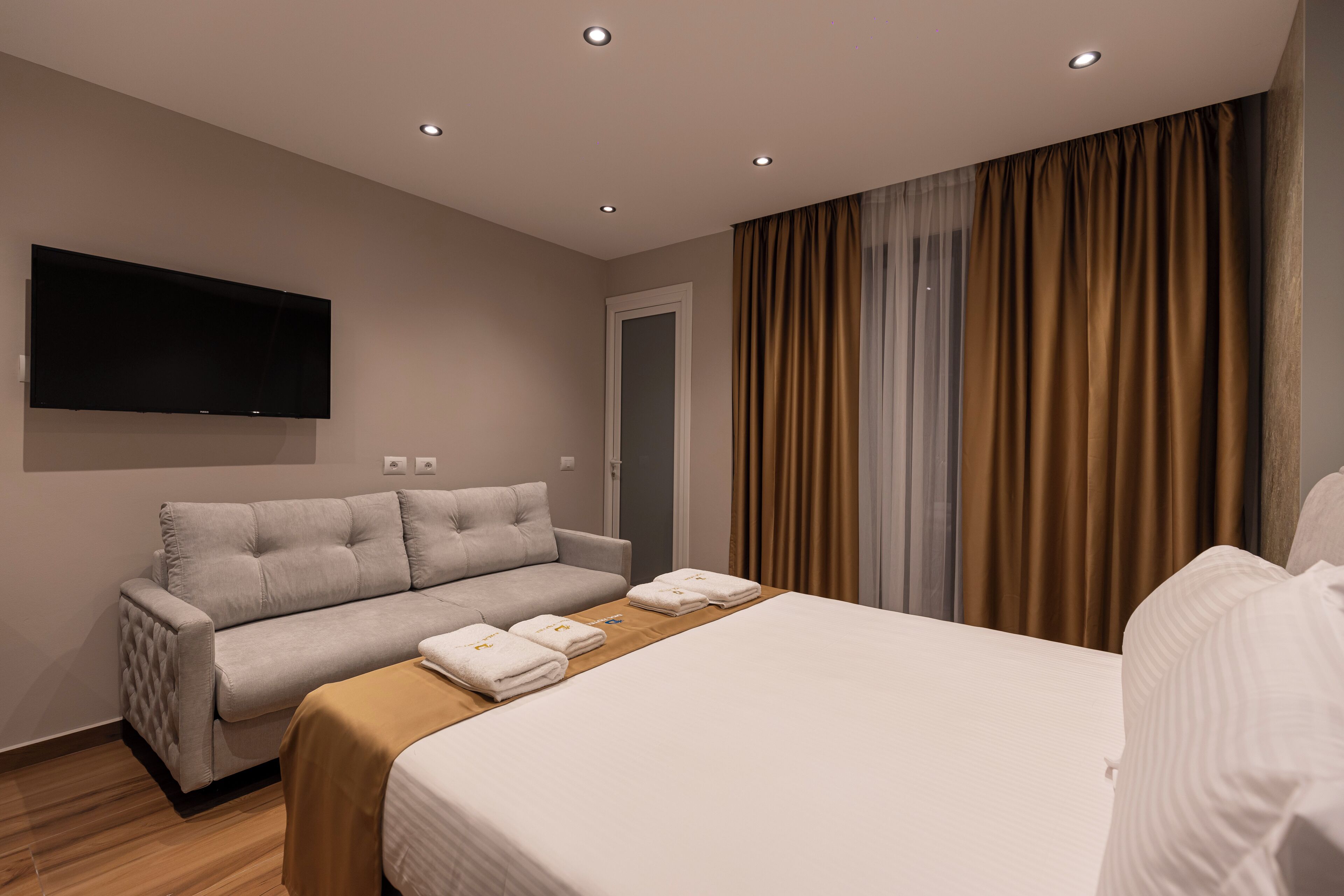 Una habitación de hotel moderna con cama, sofá, televisor de pantalla plana montado en la pared y ventana con cortinas.