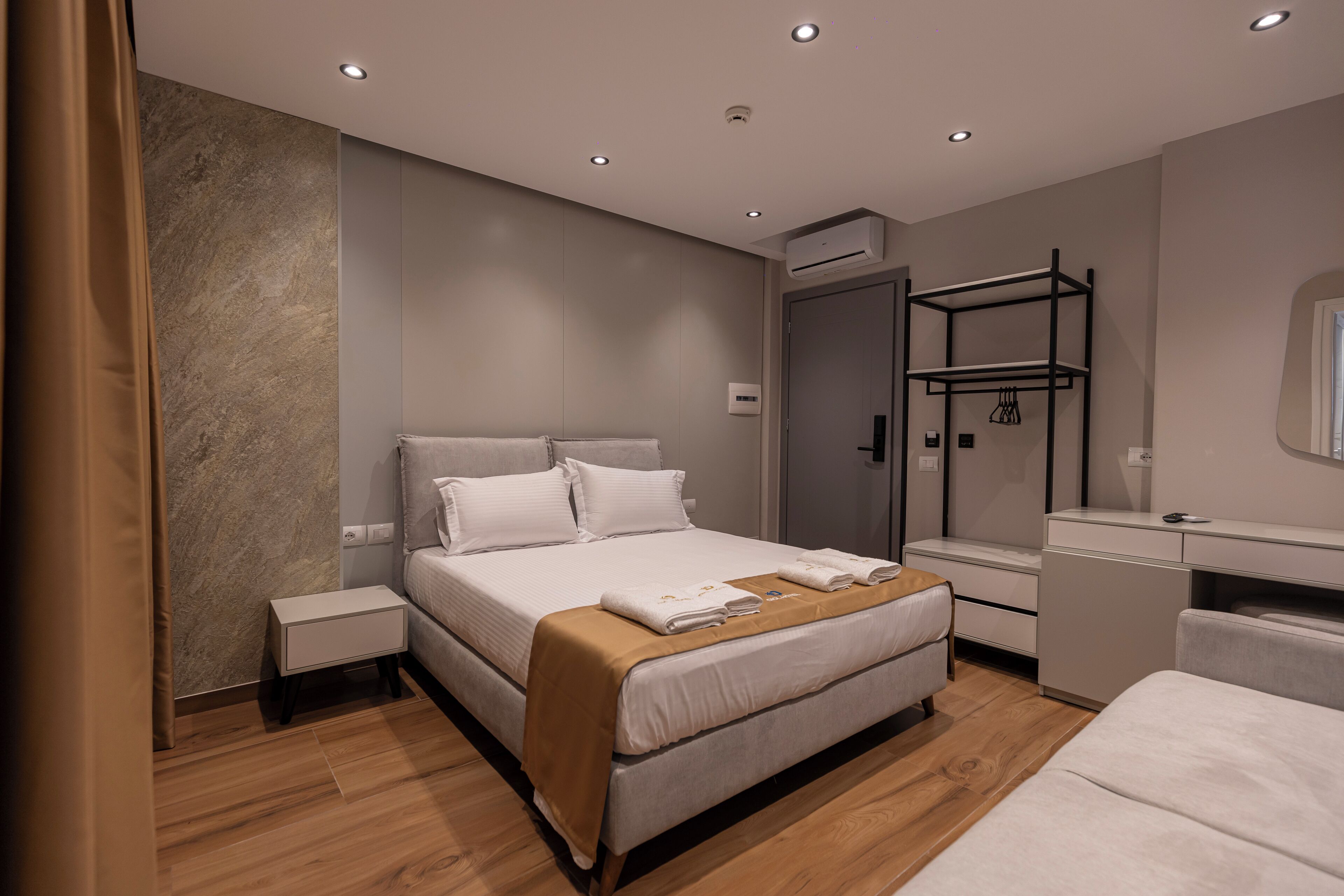 A modern hotel room with a large bed, a bedside table, a wardrobe, a desk, and a mirror.
