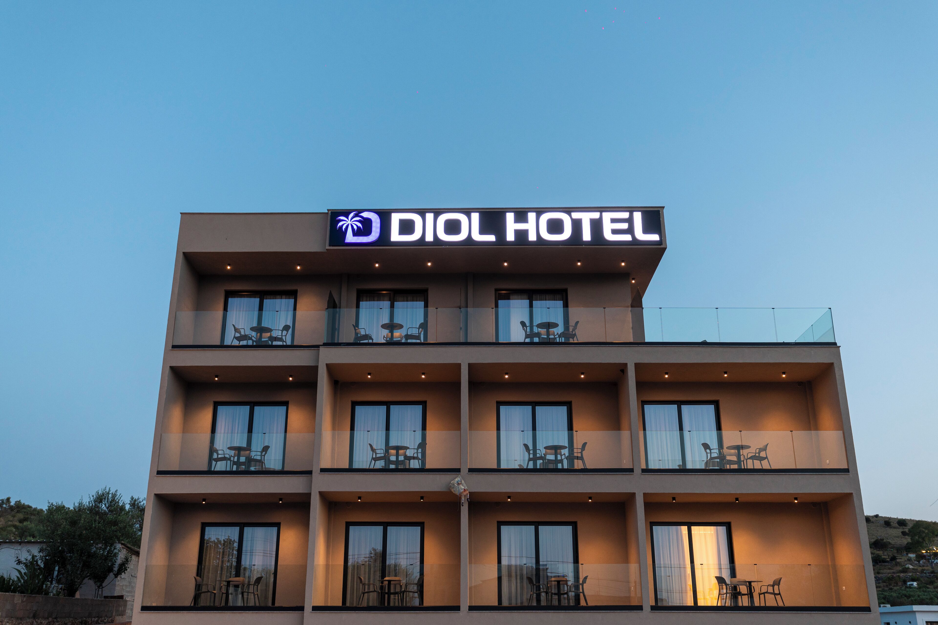 A modern hotel building with a sign reading "DIOL HOTEL". It features multiple balconies with outdoor seating and large windows.