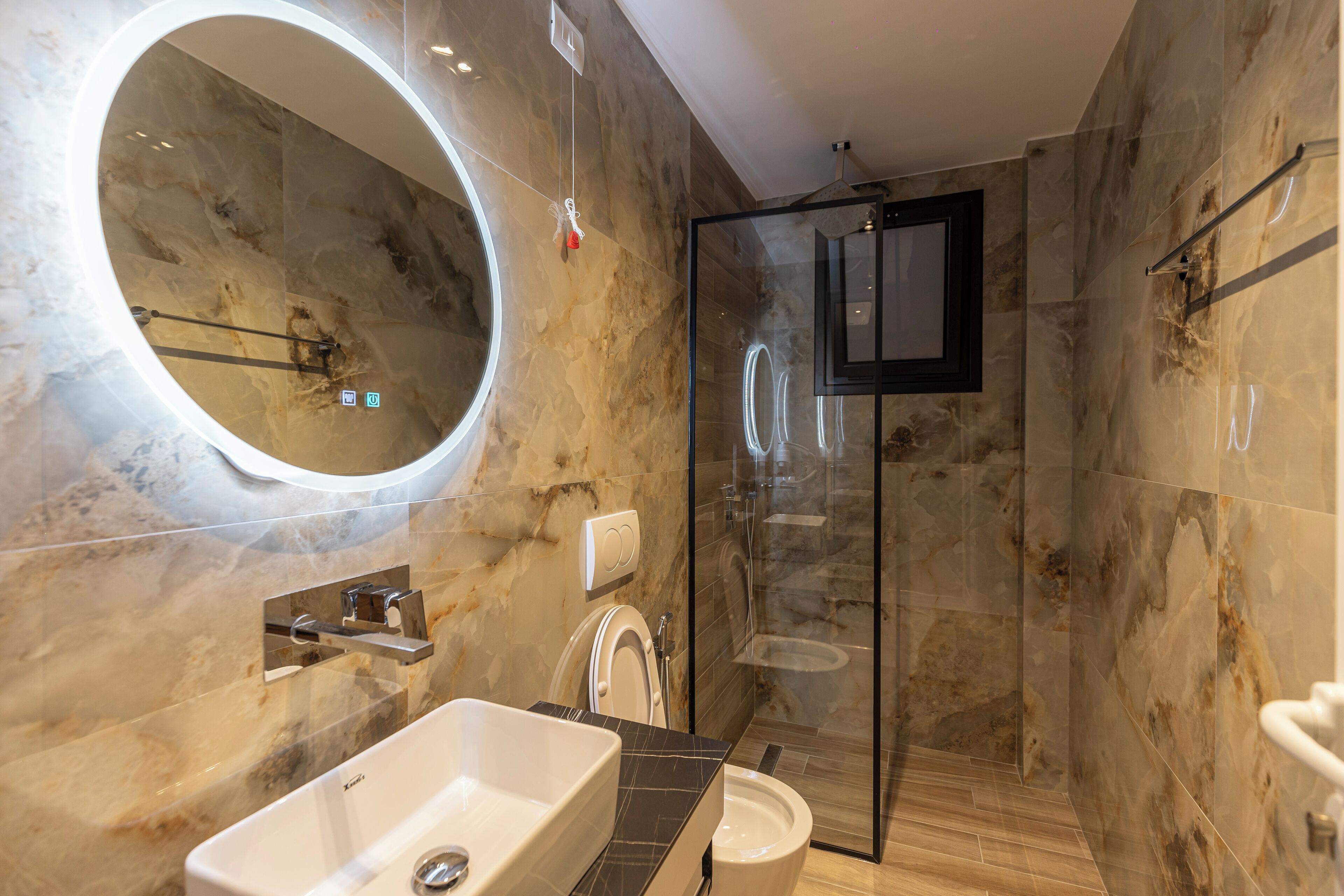 A modern bathroom with a large oval mirror, a freestanding sink, a glass-enclosed shower, and marble walls.