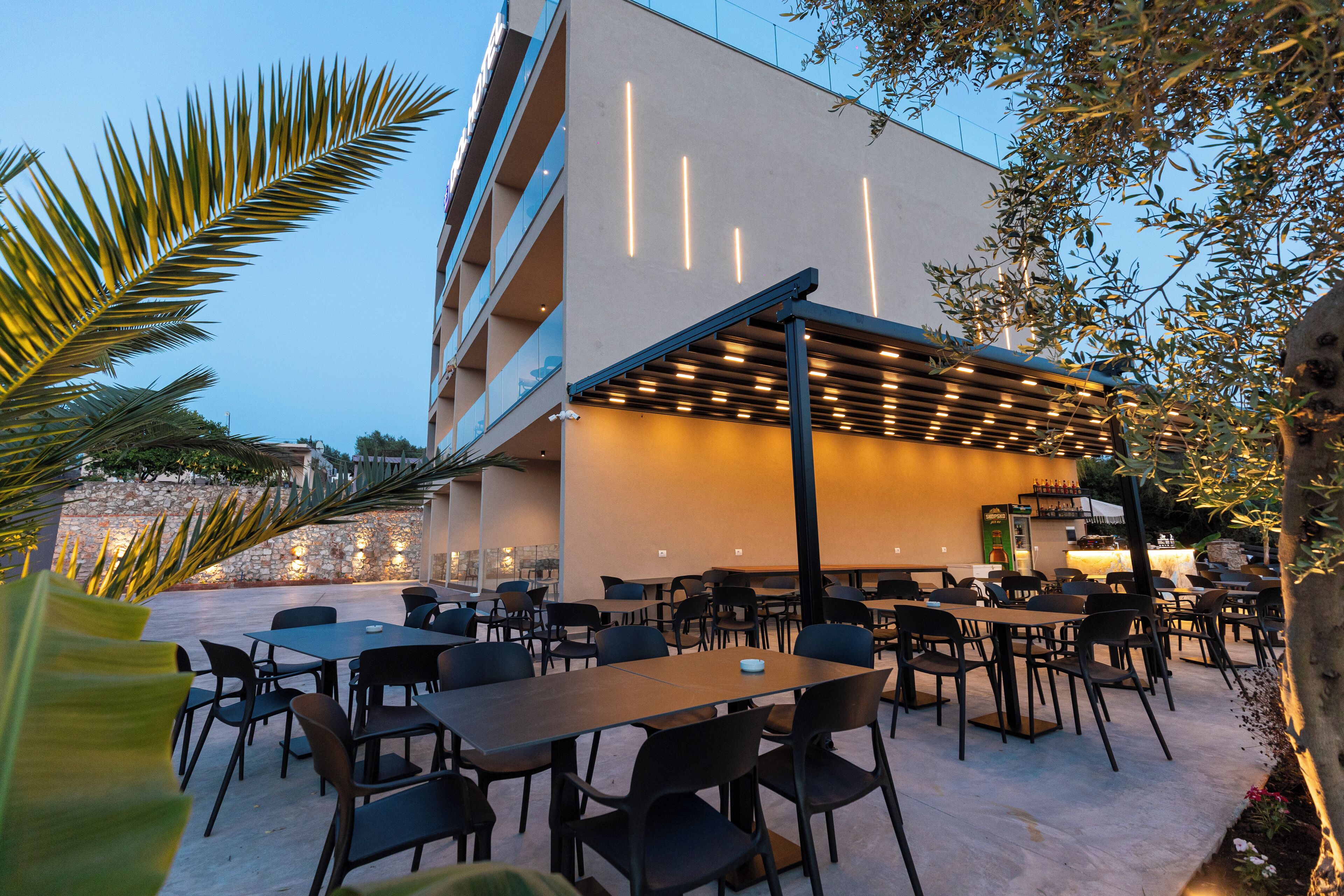 An outdoor dining area with tables and chairs, a pergola, and a building with vertical light fixtures.