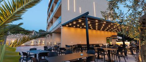 An outdoor dining area with tables and chairs, a pergola, and a building with vertical light fixtures.
