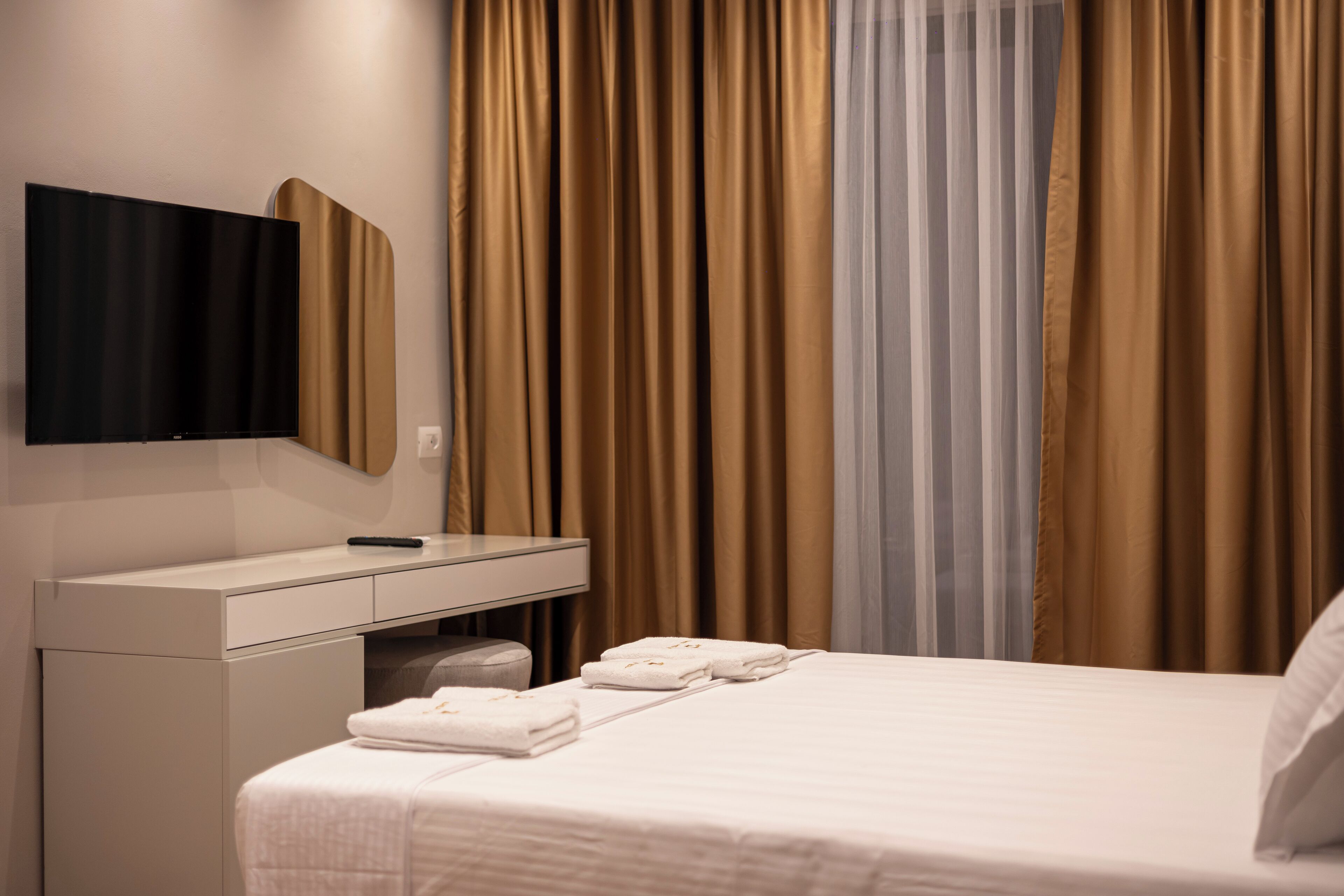 A hotel room with a bed, a desk, a flat-screen TV mounted on the wall, and a window with curtains.