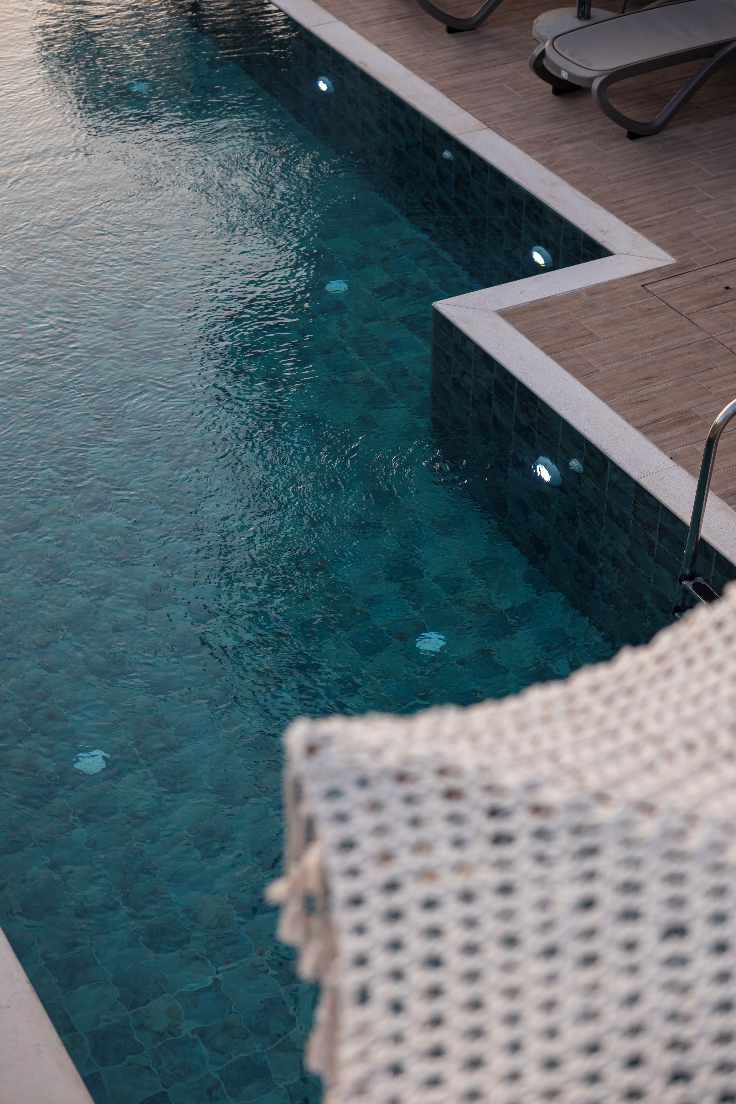 Una piscina limpida con bordo piastrellato e scaletta in metallo. Sono presenti lettini e un asciugamano bianco con trama sul bordo piscina.