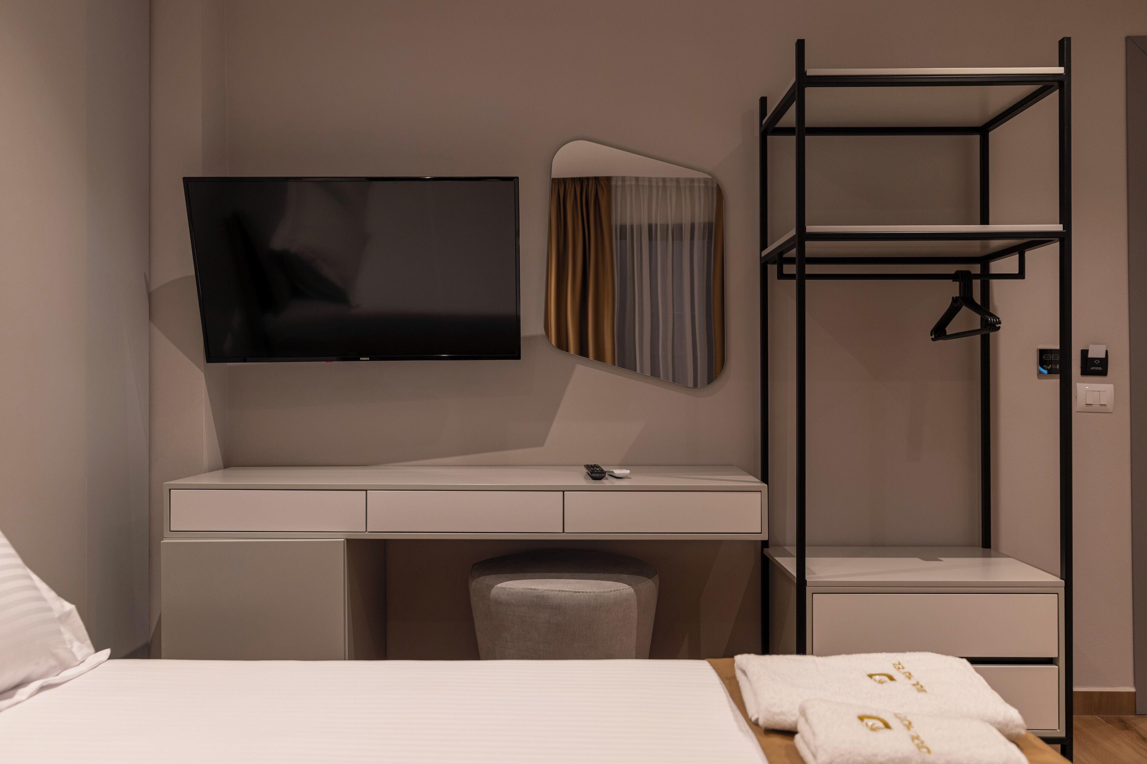 A modern hotel room with a flat-screen TV mounted on the wall, a desk with a chair, a hanging wardrobe, and a bed with neatly folded towels.