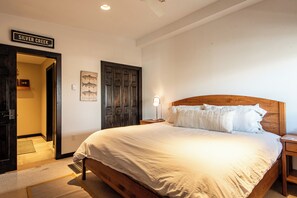 A bedroom with a large bed, a wooden headboard, a nightstand, a wall-mounted lamp, and a door labeled "SILVER CREEK".