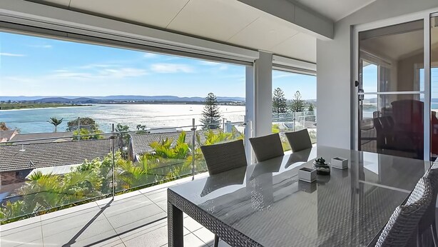 A modern dining area with a glass table, chairs, and a view of a lake and mountains. - The Sea Serenade - Coastal haven with panoramic ocean views! (Swansea Heads)